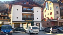 A three-story building with white and brown exterior, balconies on the second floor, and a name board 'TEA-ROOM ALETSCH'. There are several cars parked in front.