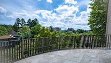 The image shows an outdoor terrace or balcony with a metal railing overlooking a scenic landscape. The terrace has a stone or concrete floor, and the view beyond the railing includes lush green trees, a building in the distance, and a partly cloudy blue s
