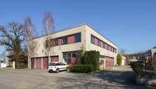 Industrial building with red garage doors, white car parked in front, large windows, green bushes, clear sky