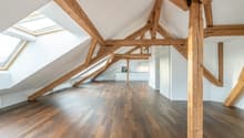 Spacious attic room with exposed wooden beams, hardwood flooring, and a skylight providing natural light. The room appears to be an open-concept living space with a kitchen visible in the background.