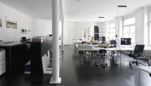Large open office space with multiple desks, computers, and office equipment. The room has high ceilings, white walls, and dark hardwood floors. There is a dishwasher visible in the corner of the room.