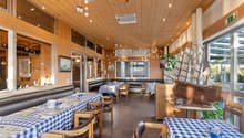 Cozy wooden interior with checkered tablecloths, leather seating, and nautical decor elements like a model ship. The space features pendant lighting, an air conditioning unit, and a dishwasher visible in the background.