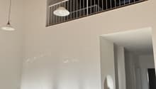 The image shows the interior of a room with a high ceiling and exposed wooden beams. There is a balcony or terrace visible through the railing on the upper level. The room has a minimalist design with white walls and a wooden bench or shelf below the balc