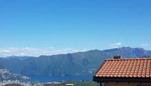 The image shows a scenic view of a mountainous landscape with a clear blue sky. In the foreground, there are rooftops of buildings with red tiles. The background features mountains and a body of water, likely a lake or a bay.