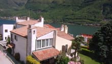 Large house with red roof, white walls, multiple windows, surrounded by greenery, mountains in the background, lake nearby