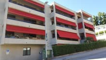Multi-story apartment building with red and white exterior, balconies on each floor, and a parking area in front of the building.