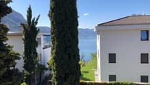 Two white buildings, one with a balcony, surrounded by greenery, with a lake and mountains in the background.