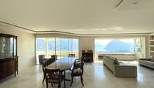 Spacious room with a dining table, chairs, a cabinet, a couch, an ottoman, a fireplace, and bookshelves. Large windows offer a scenic view.