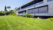 A modern gray building with large windows and a glass balcony. It has a spacious lawn with bushes and trees in front.