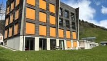 A modern building with gray and orange exterior, raised ground floor, large glass windows, surrounded by green grass and a car parked outside.
