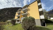 Yellow building with balconies on each floor, stairs leading up to building, small plants and bushes in front