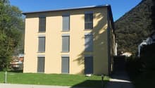 Modern yellow building with 8 windows and a flat roof, with grass and trees around, located on a raised ground floor
