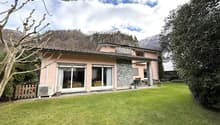 Detached house, pink exterior, large windows, spacious lawn, exterior AC unit, patio with chairs, trees, bushes, mountain in the background