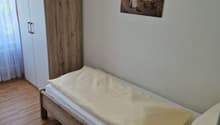 The image shows a bedroom with a wooden wardrobe, a bed with white bedding, and a framed artwork on the wall. The room has a hardwood floor and appears to be a simple, functional living space.
