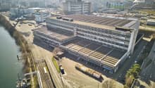 Large multi-story industrial building with a flat roof, surrounded by other commercial and industrial buildings. The building has a rectangular shape and appears to be used for manufacturing or warehousing purposes. There are several vehicles parked in th
