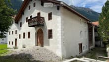 Large white building with brown roof, 2 floors, many windows, wooden door, wooden balcony, gravel path, fountain