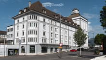 Multi-story building, white exterior, brown roof, many windows, shops on the ground floor, parking lot in front