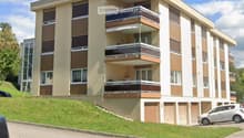 A three-story residential building with balconies, white walls, brown frames, underground parking, and a grassy hill in front.