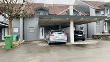 grey residential building with tiled roof, covered parking lot, two cars parked, two trash bins, tree, small stone wall