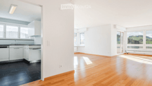 Open-plan space with wooden flooring, white walls, and large windows. The area includes a kitchen section with cabinets and a sink, and a living area with ample natural light.