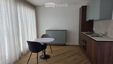 Modern kitchen with wooden floor, white walls, and gray cabinets. Includes a round white table, two chairs, and a sink with a faucet. A power outlet is mounted on the wall.
