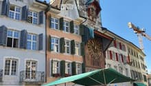 Town square with buildings, large clock tower, green market umbrellas, plants for sale