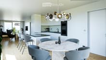 Marble table, round shape, modern chairs, central chandelier, white cabinets, black countertop, kitchen area with oven, modern furniture, wooden floor, windows with curtains