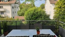 Outdoor table with 4 chairs, balcony, black and white cushions, plants, trees, view of buildings