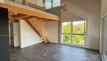 Open-plan living space with exposed wooden beams, grey tiled floor, large windows, and a wooden staircase leading to an upper level.
