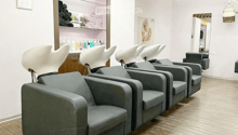 A salon with a row of grey chairs, a wooden floor, a white wall, a shelf with products, and a mirror.