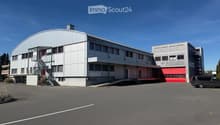Large industrial building with white walls, red doors, and a large parking area in front