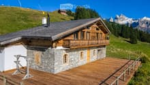 A two-story wooden chalet with stone base, large balcony, and wooden deck, surrounded by green hills and mountains.