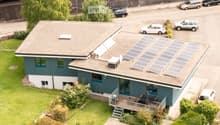 Blue house with solar panels on the roof, wooden deck with outdoor seating, multiple cars parked in the parking lot