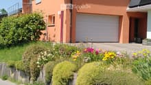 Two-story house with orange exterior, white garage door, satellite dish, and well-maintained garden with colorful flowers and shrubs.