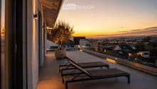 Modern rooftop terrace with sun loungers, a small table, and a potted tree, overlooking a cityscape at sunset.