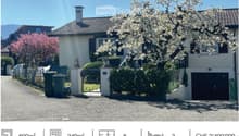 A two-story house with a tiled roof, white exterior walls, and black shutters. The house is surrounded by a garden with a variety of plants and trees, including a cherry blossom tree in full bloom. The driveway is paved with gravel, and there is a black g