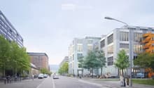 Urban street scene with modern buildings, streetlights, and trees. A few people are walking on the sidewalk, and some bicycles are parked. Vehicles are driving on the road.