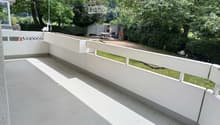 A modern, spacious balcony with white railings and a concrete floor, overlooking a lush green garden with trees and a small seating area.