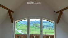 Empty room with wooden floor, white walls, and large glass doors with wooden frames. View of a green field and mountains.