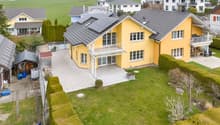 Aerial view of a two-story yellow house with a grey roof, surrounded by a well-maintained lawn, garden, and neighboring houses in a suburban setting.