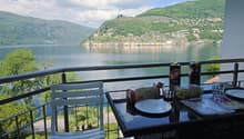 Balcony / Terrace. Balcony with a scenic lake view, featuring a table with chairs, a bottle, bread, and a cup.