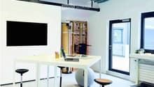 Furnished Room. Modern office space with white tables, laptop, stools, a large screen, a ball chair, and a treadmill.