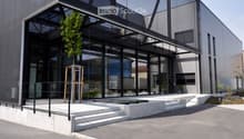 Building. Modern building with large glass windows and a sleek, contemporary design. Features a raised ground floor, small water feature, and small landscaped area with plants and trees.