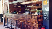 Wooden bar counter with hanging wine glasses, bottles, and stools, illuminated by hanging lamps.