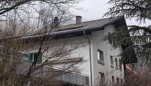 House. A two-story house with white walls, multiple windows, and a tiled roof, partially obscured by trees and branches.