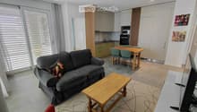 Furnished Room. Open-plan living and dining area with a grey sofa, wooden coffee table, dining table with chairs, and a small kitchen area with cabinets and appliances.