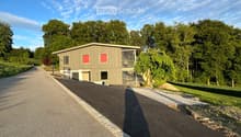 Building. A modern, single-story building with a flat roof and red-framed windows, situated on a paved driveway surrounded by greenery.