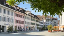 Garden / Outdoor Area. A European town square with colorful buildings, outdoor seating, and trees. The square is paved and features potted plants and umbrellas.