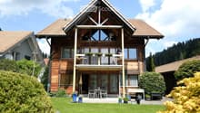 House. Two-story wooden house with a triangular roof, large windows, and a balcony. The house is surrounded by a well-maintained garden with a patio area, outdoor furniture, and potted plants.