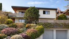 Two-story house with a raised ground floor, modern design, large windows, and a balcony on the upper level. The house features a garage and is surrounded by well-maintained gardens and trees.